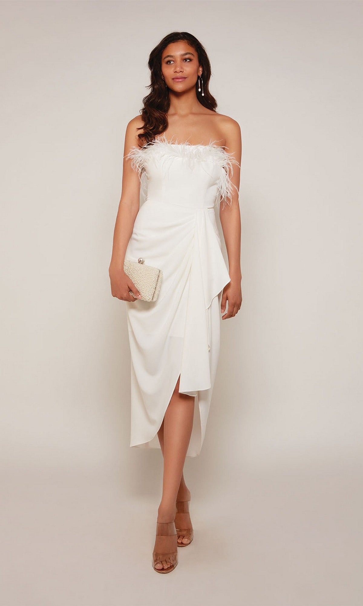 A woman poses against a plain background in the Alyce 70033 knee-length, white off-the-shoulder semi-formal dress with feather accents, styled with a small clutch and nude heels.